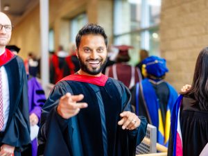 A faculty member in a Convocation gown smiles with his hands up pointing forward.