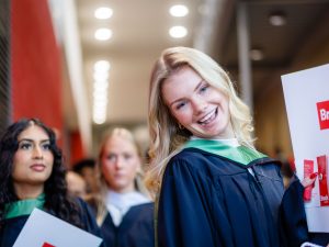 A woman in a graduation gown smiles while holding up a folder with Brock University on the front.