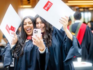 Two women in Convocation gowns take a selfie together while each holding up folders with Brock University on them.
