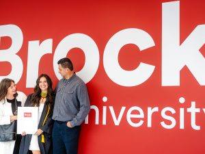 Three people, one in a graduation gown, stand against a wall with a large Brock University logo.