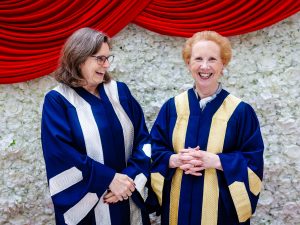 Two women in Convocation gowns stand together against a wall of white flowers.