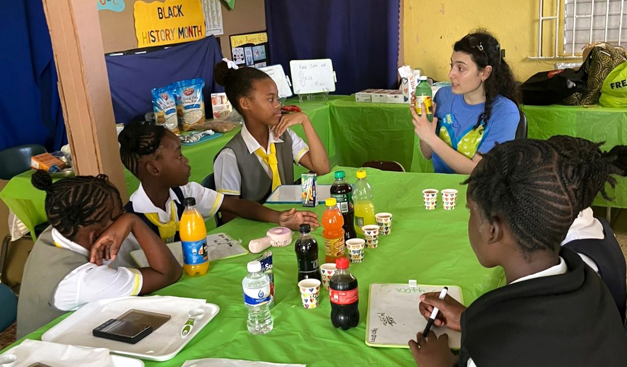 There is a woman in a blue and green shirt showing the label affixed to a bottled drink while children gathered around the table listen.