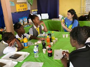 There is a woman in a blue and green shirt showing the label affixed to a bottled drink while children gathered around the table listen.
