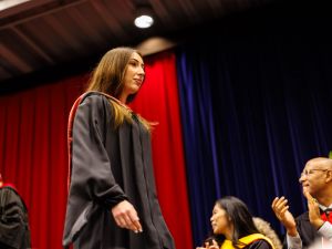 A young woman in academic robes walks across the stage during a university graduation ceremony.