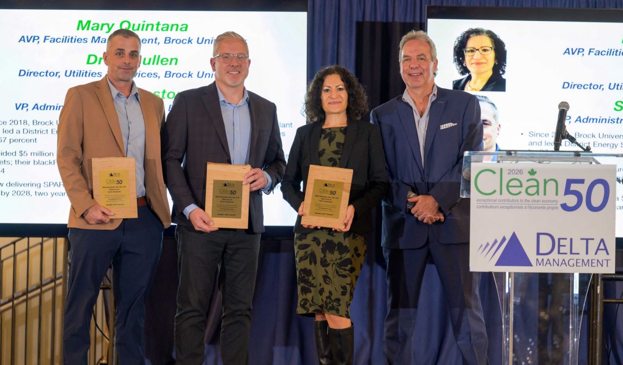 Four people stand on a stage at the Clean50 awards ceremony, with three award recipients holding wooden plaques while posing beside a presenter. Large presentation screens and a Clean50 podium are visible in the background.