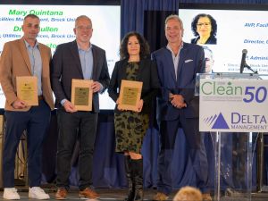Four people stand on a stage at the Clean50 awards ceremony, with three award recipients holding wooden plaques while posing beside a presenter. Large presentation screens and a Clean50 podium are visible in the background.
