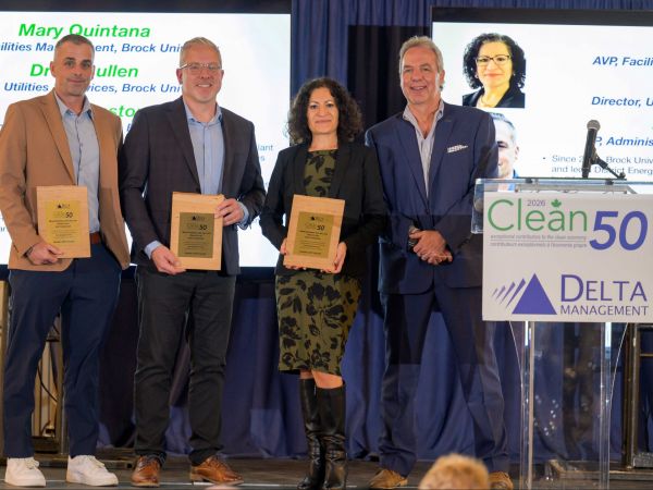 Four people stand on a stage at the Clean50 awards ceremony, with three award recipients holding wooden plaques while posing beside a presenter. Large presentation screens and a Clean50 podium are visible in the background.
