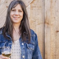 "Leanne Molchan smiling in a denim jacket, holding a glass of wine against a rustic, light-wood barn wall."