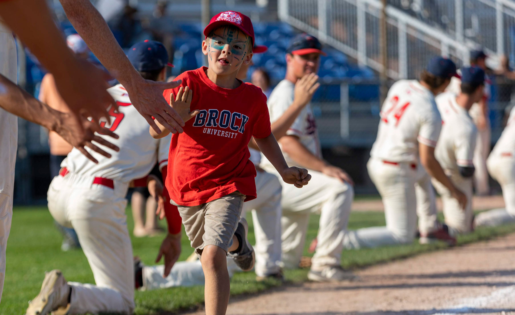 Young boy wearing a Brock University t-shirt and face paint running and high-fiving members of the Brock men's baseball team