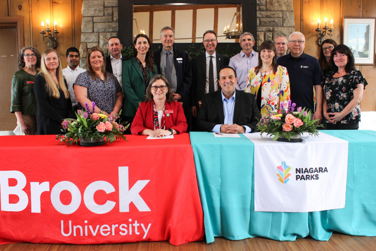 Representatives from Brock University and Niagara Parks stand behind MOU Signatories Dr. Lesley Rigg and Graham Coveney, who sit behind tables branded with Brock University and Niagara Parks logos. 