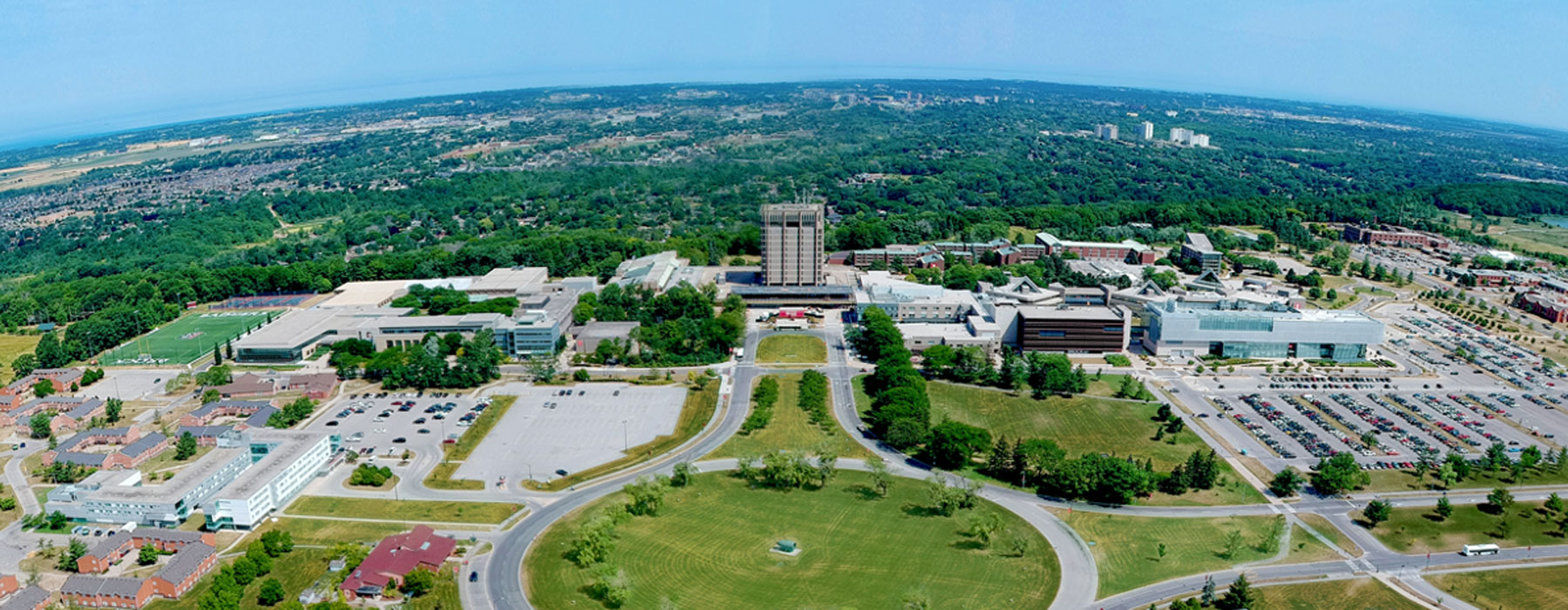 An aerial view of Brock's campus. The green lawn of the entrance is in the foreground, leading to Schmon tower in the midground and horizon, with a clear blue sky. 