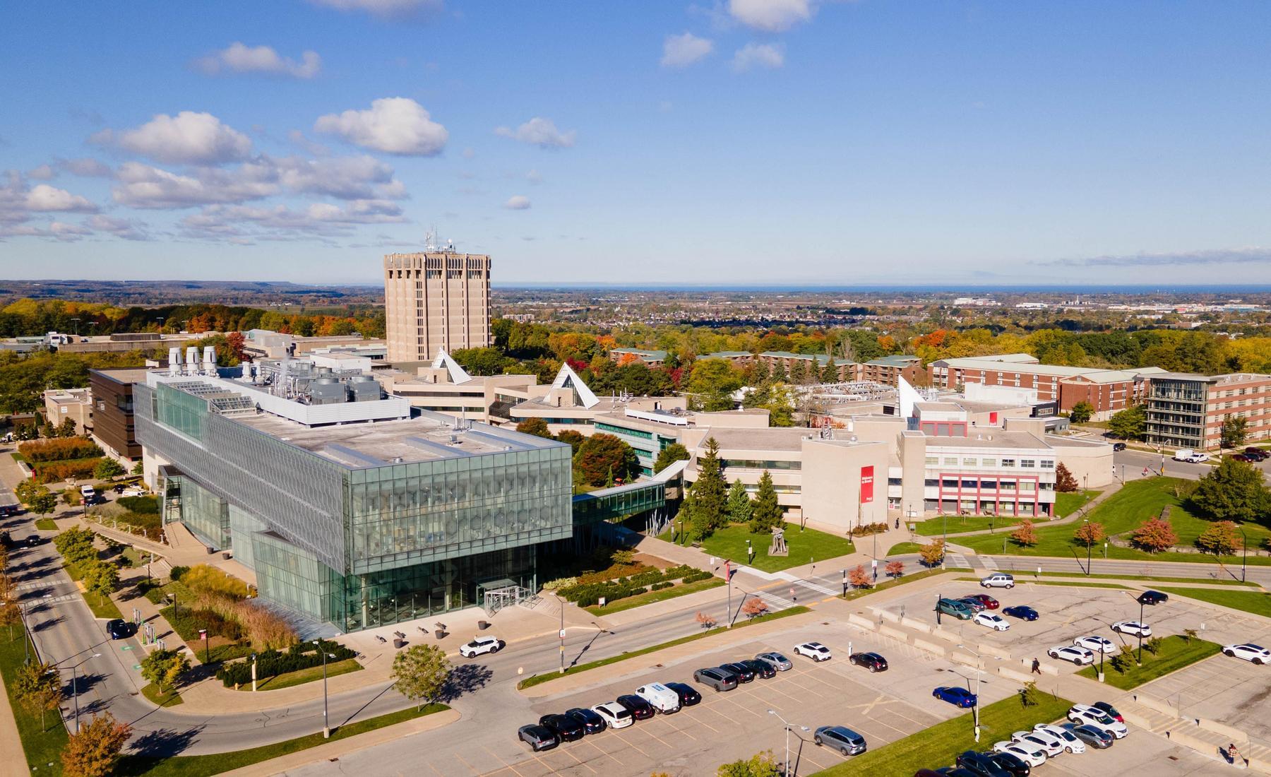 Aerial image of Brock University's main campus. Parking lot in foreground. Campus buildings surrounded by natural landscape in background. 
