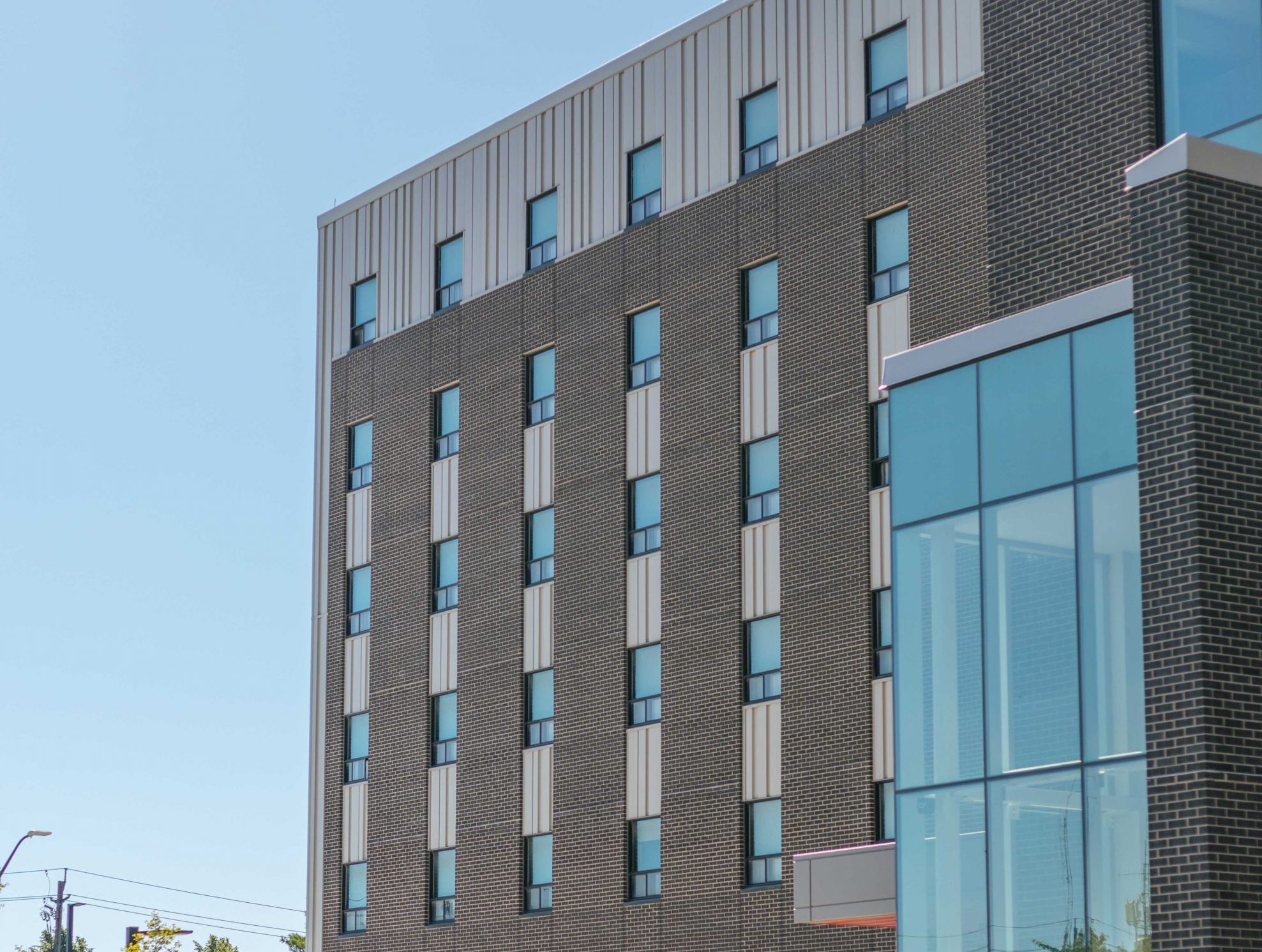 Residence 8 Exterior - Compressed Exterior of Residence 8 at Brock University, featuring a modern brick and glass façade with tall windows, landscaped greenery, and a clear blue sky.
