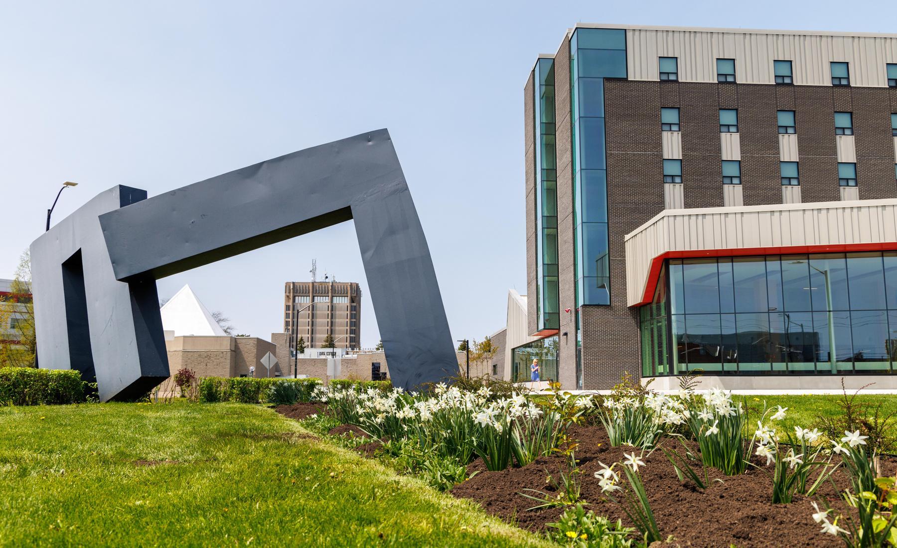 Image of Brock University's campus. Garden, abstract artistic sculpture in left foreground. Residence 8 building in right foreground. Schmon Tower and other campus buildings in background. 