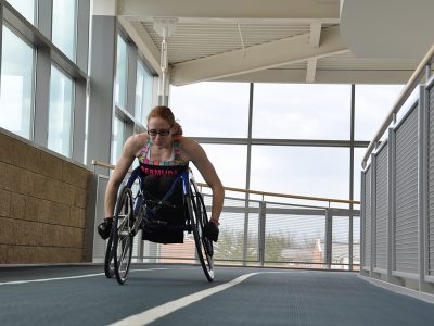 woman in a wheel chair on the 200m indoor track is suspended above the Ian Beddis gymnasium