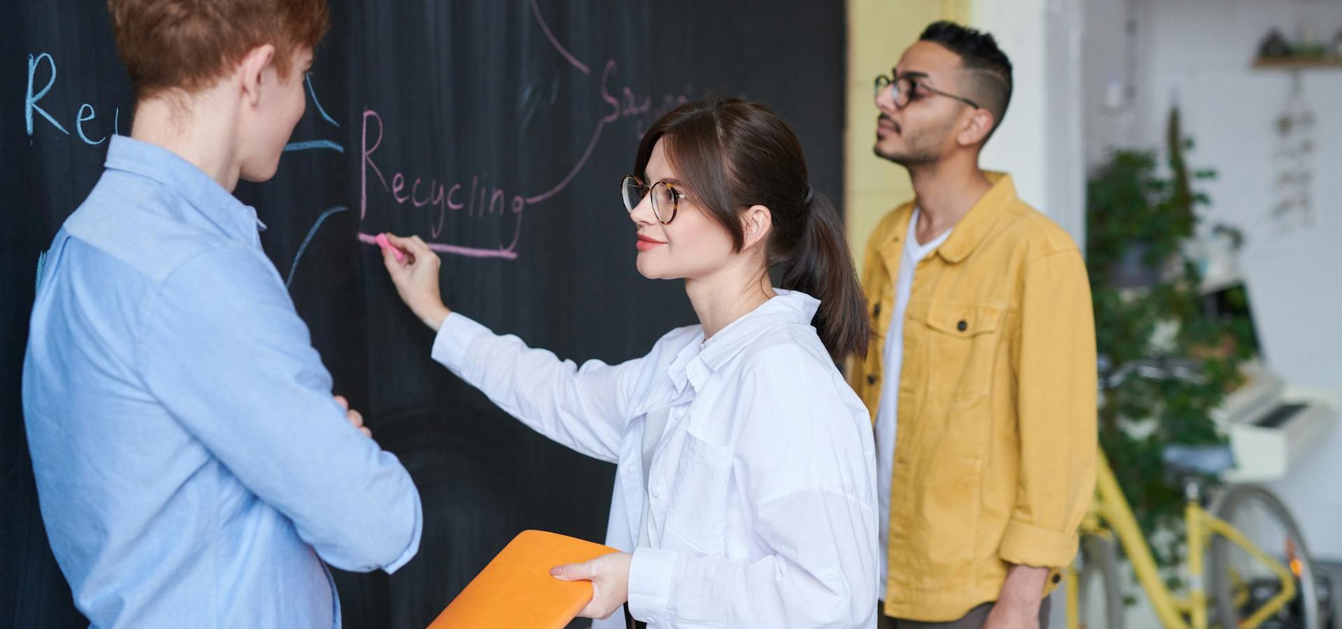 3 people staring at a chalkboard with a woman writing the word "recycling" on the chalkboard
