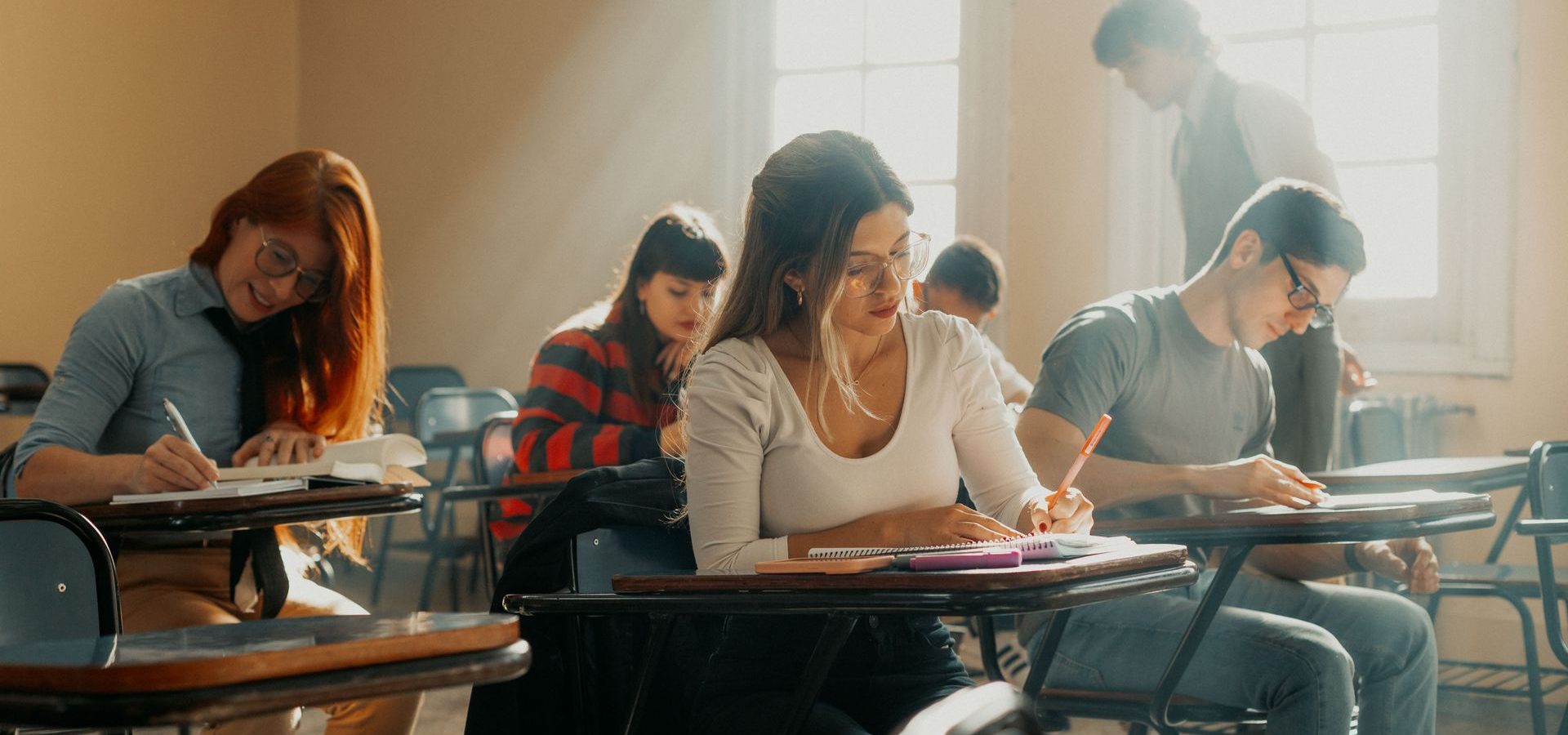 Students sitting at desk writing on paper as teacher watches over them