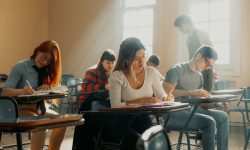 Students sitting at desk writing on paper as teacher watches over them