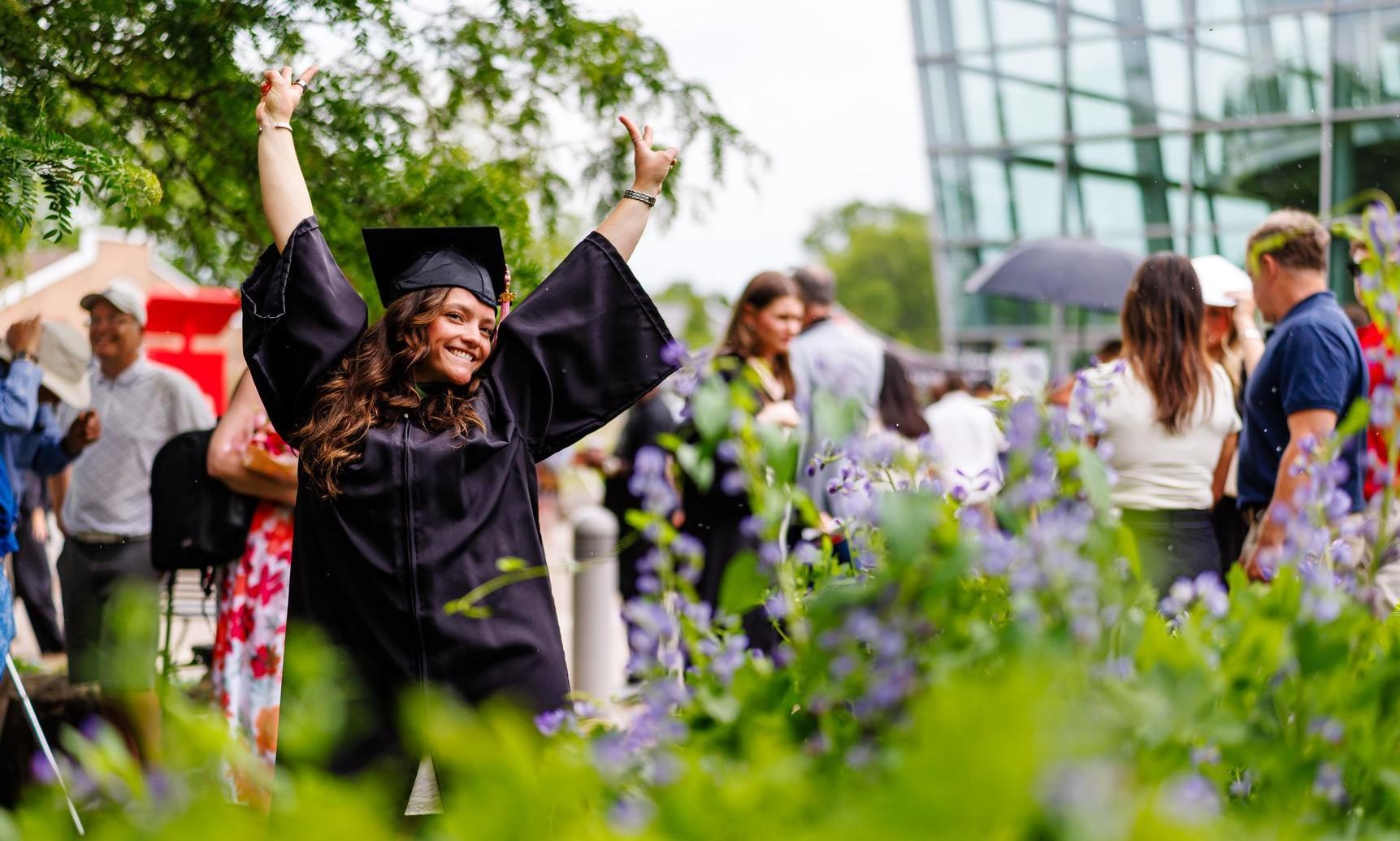 Graduand in a black academic gown and mortarboard raises both arms while standing among purple flowers outside a glass-fronted campus building, with convocation attendees gathered in the background. 