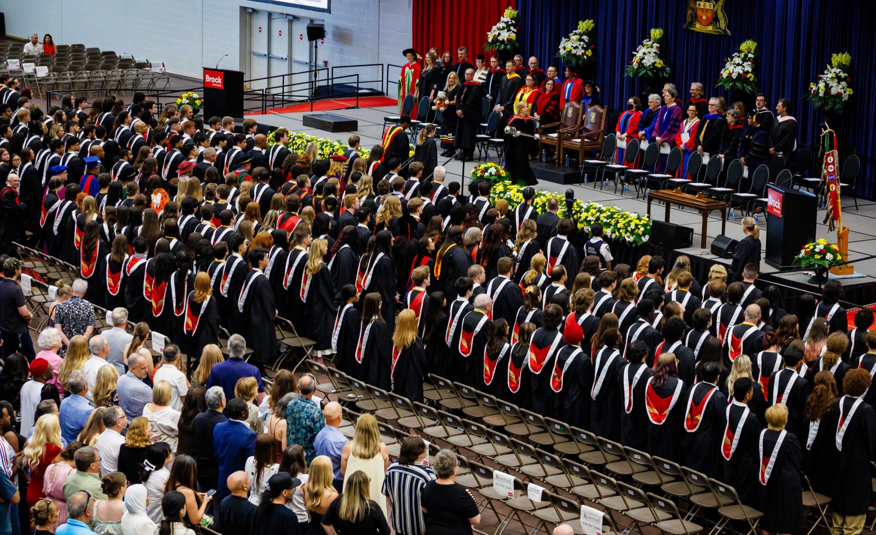 Graduates in academic regalia stand during a Brock University Convocation ceremony as faculty and university officials gather on stage.