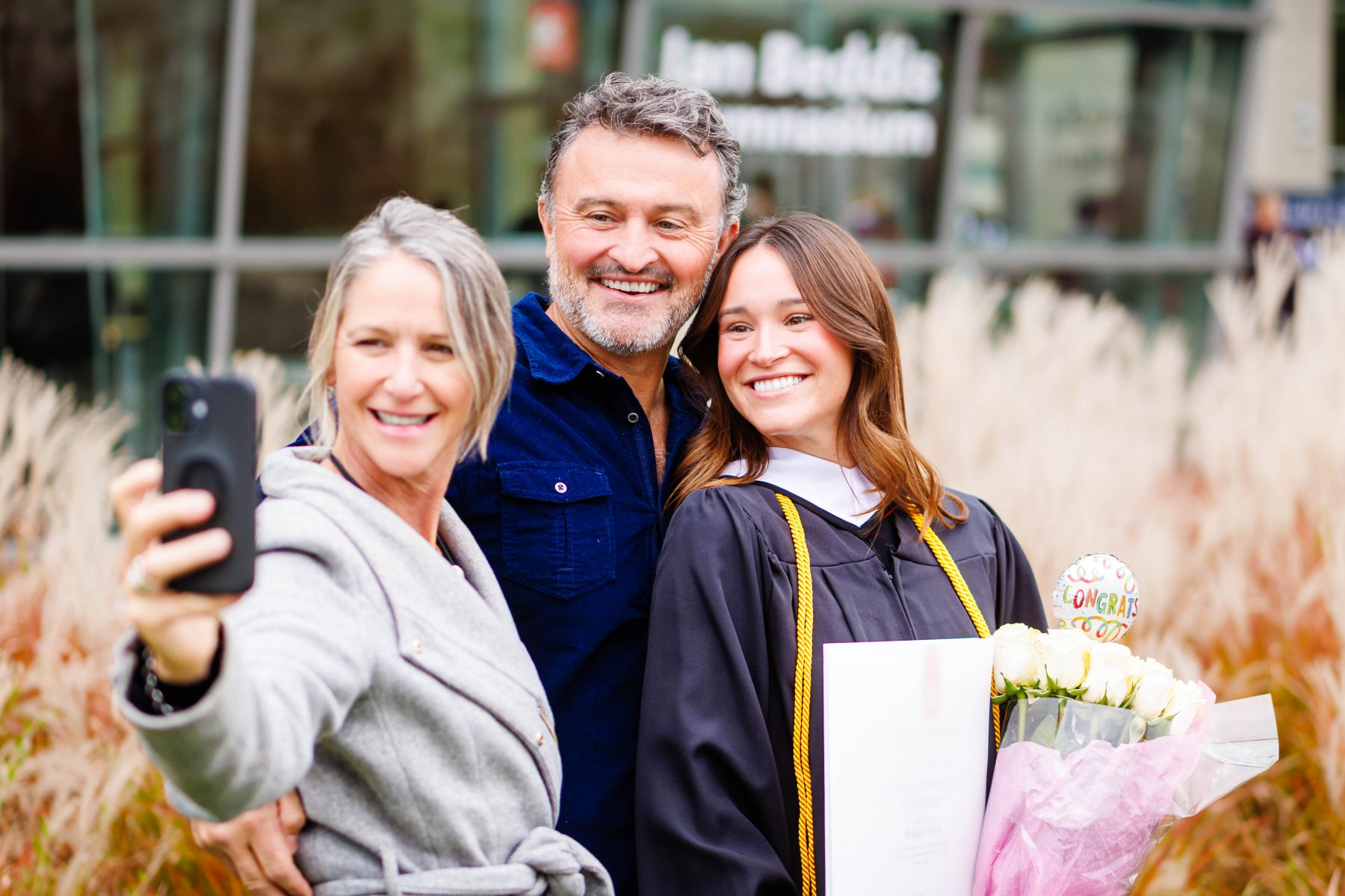 Graduate holding flowers and a diploma cover poses for a selfie with two supporters following a convocation ceremony.