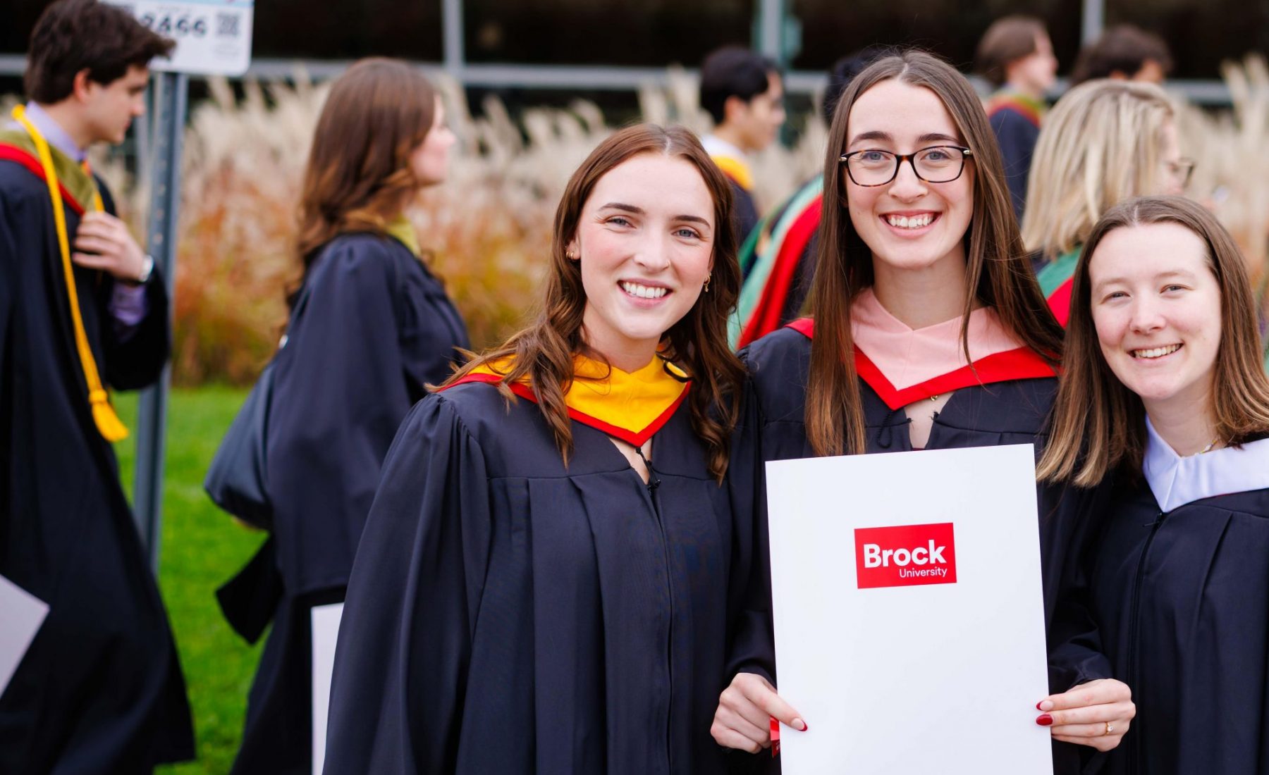 Three Brock University graduates wearing academic regalia stand together on a lawn during convocation and face the camera. The graduate in the centre holds a white diploma folder displaying the Brock University logo. Additional graduates in gowns are gathered in the background outside a campus building.