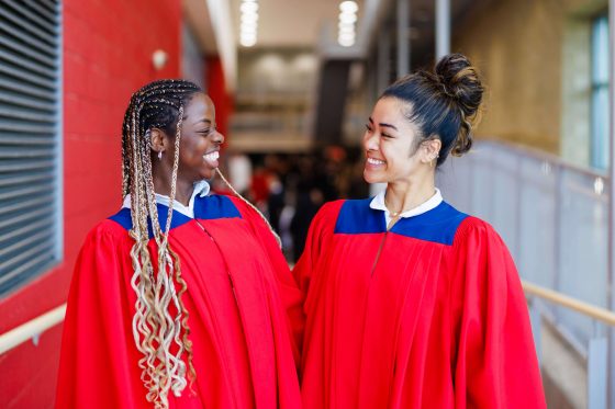 FallConvoMorning_54 compressed Two Brock University student volunteers in red Convocation gowns standing together in a campus building.