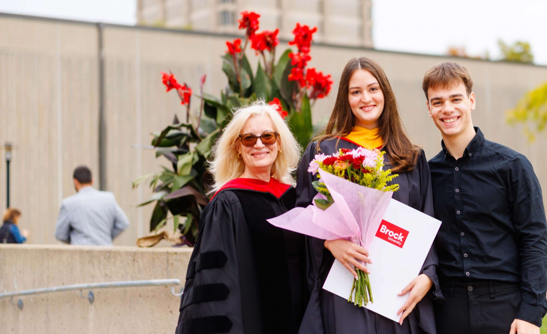 Graduate in academic regalia holding a bouquet and diploma folder while posing outdoors with two guests at Brock University Convocation. 