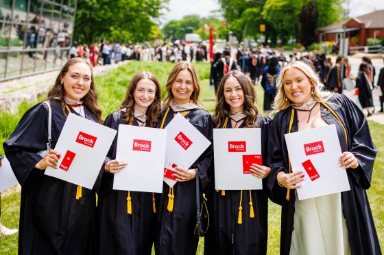 GoodmanConvocation_10 Five Brock University graduates in convocation gowns holding diploma folders outdoors following a Convocation ceremony.