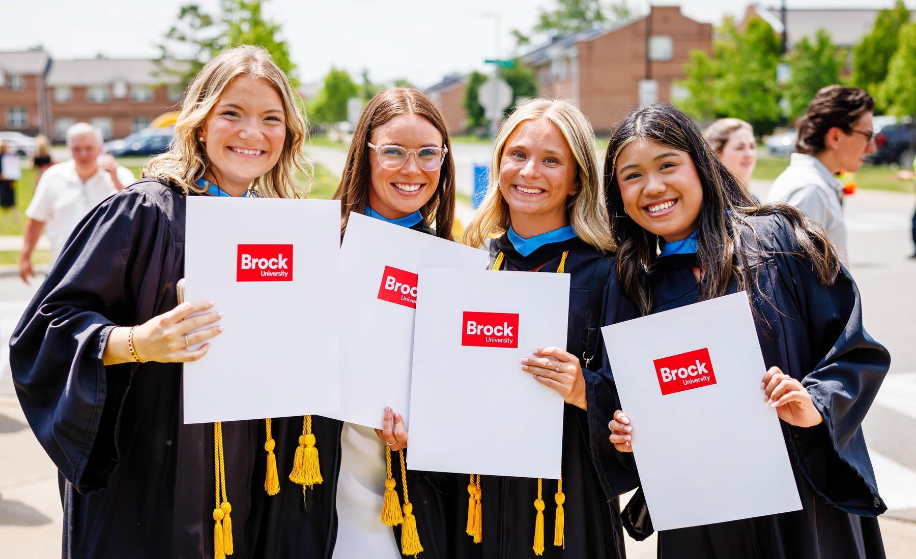 Four Brock University graduates in convocation gowns holding diploma folders outdoors on campus.