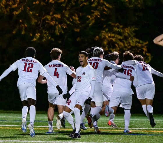 Brock's Men's Soccer Team celebrating by running into a huddle together, one player is facing the opposing team with a big celebratory expression.