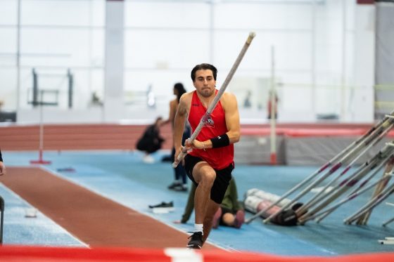 An athlete inside a gym running to practice his high jump, holding the pole.