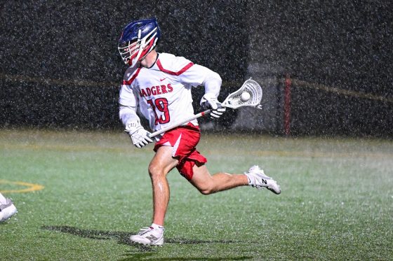 Men's Lacrosse- player in mid game running with ball, wearing a helmet. It is raining.