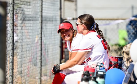 Woman's softball players sitting on a bench, one smiling at the camera.