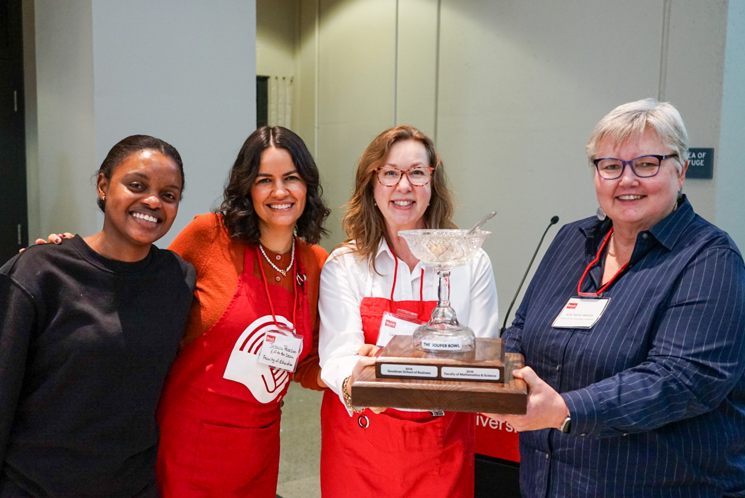 A group of four women, two of which wear a red United Way branded apron, stand together showing off a trophy shaped like a soup bowl. 