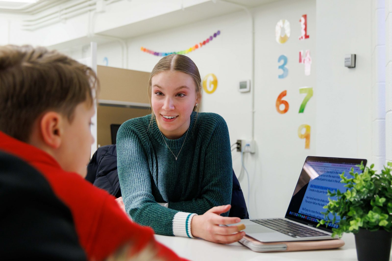 A woman sits at a desk in a classroom across from a young boy teaching on a computer.