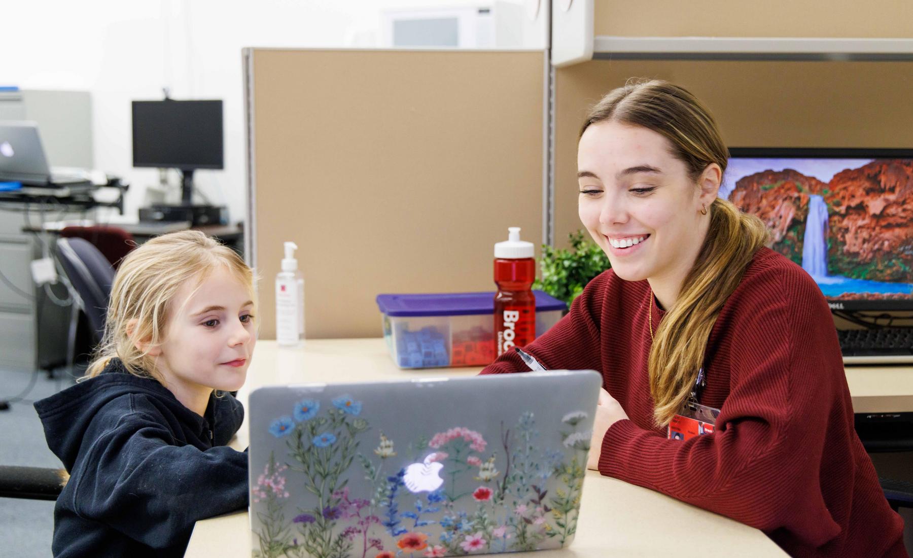 A young woman sits at a table across from a school-aged child while both look at a laptop that is decorated with colourful flower stickers. Both people are smiling warmly in the well-lit, bright room. 