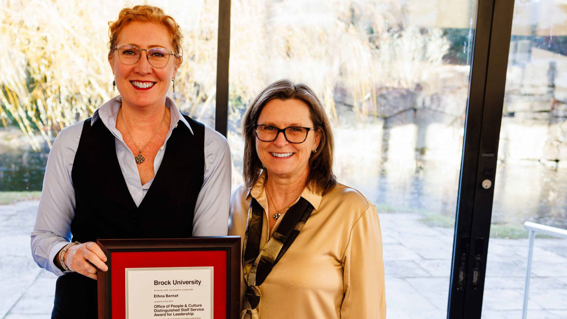 Two women stand side by side smiling, one woman holds a framed awards certificate. 