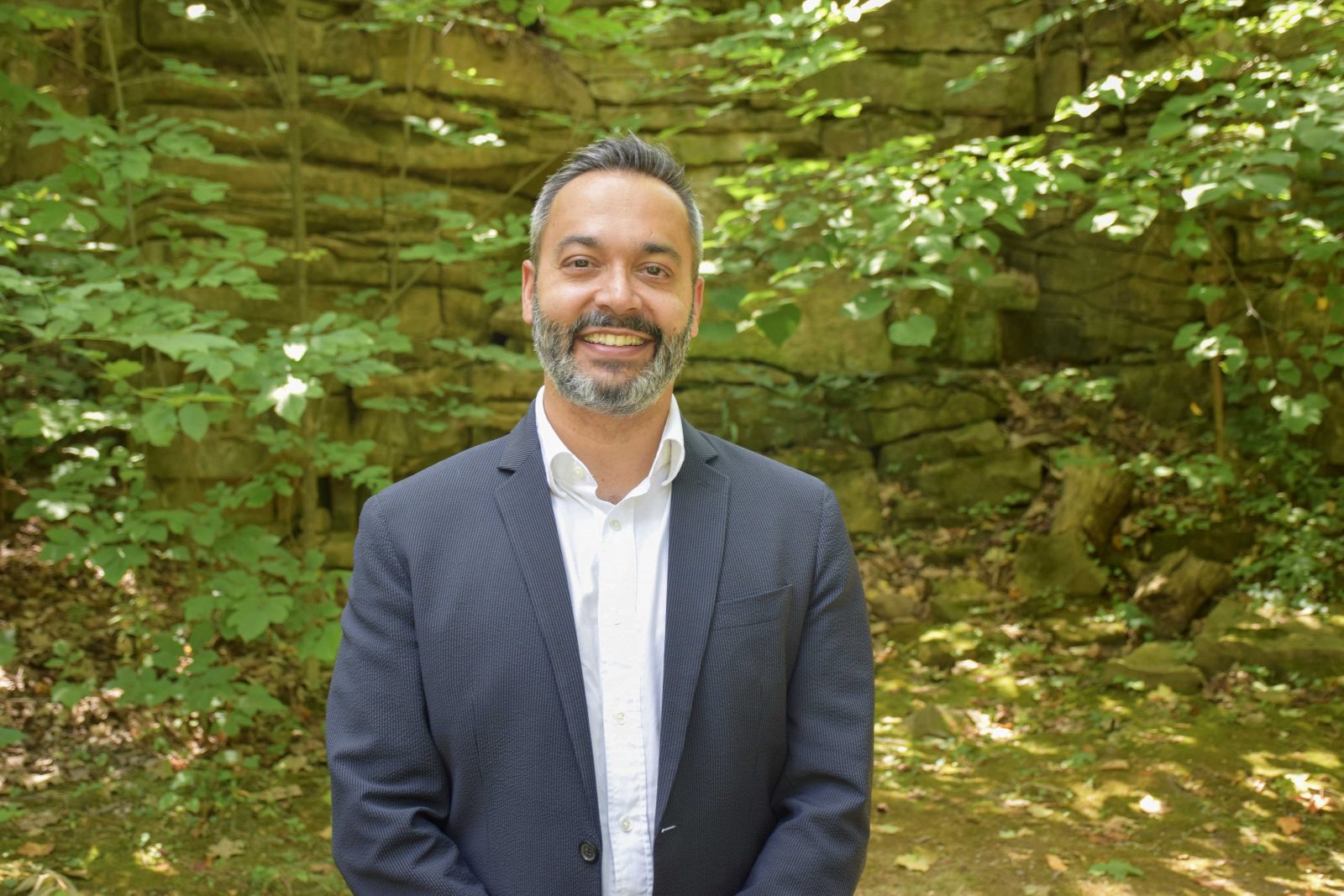 A man in a suit smiles at the camera while standing outdoors with greenery in the background. 