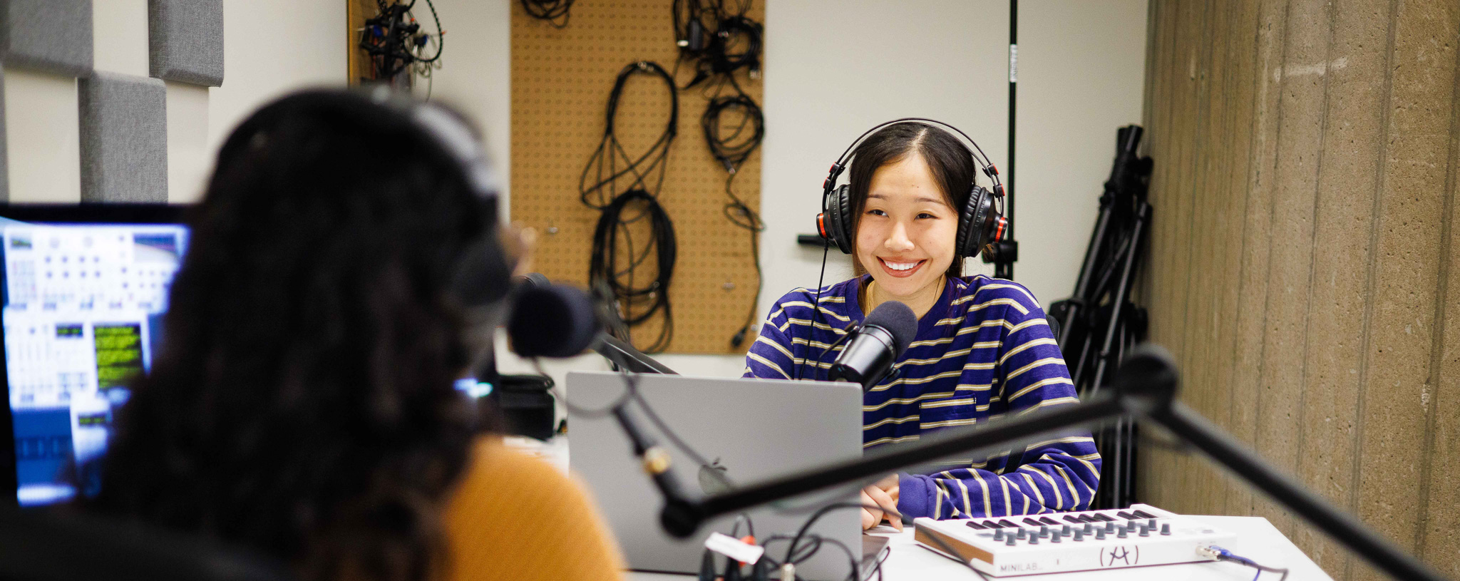 Two women sit at a desk at computers wearing headphones.