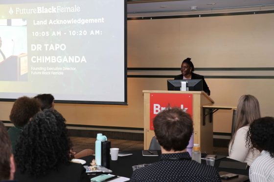 A woman stands at a podium speaking to an audience. 