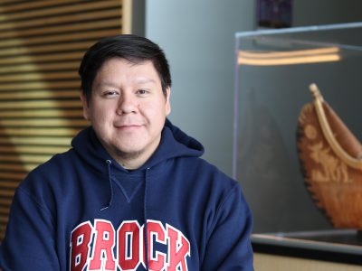 Portrait of Bobby Henry in the atrium of Welch Hall at Brock University. He is wearing a navy blue Brock sweatshirt and is sitting beside a birchbark canoe.