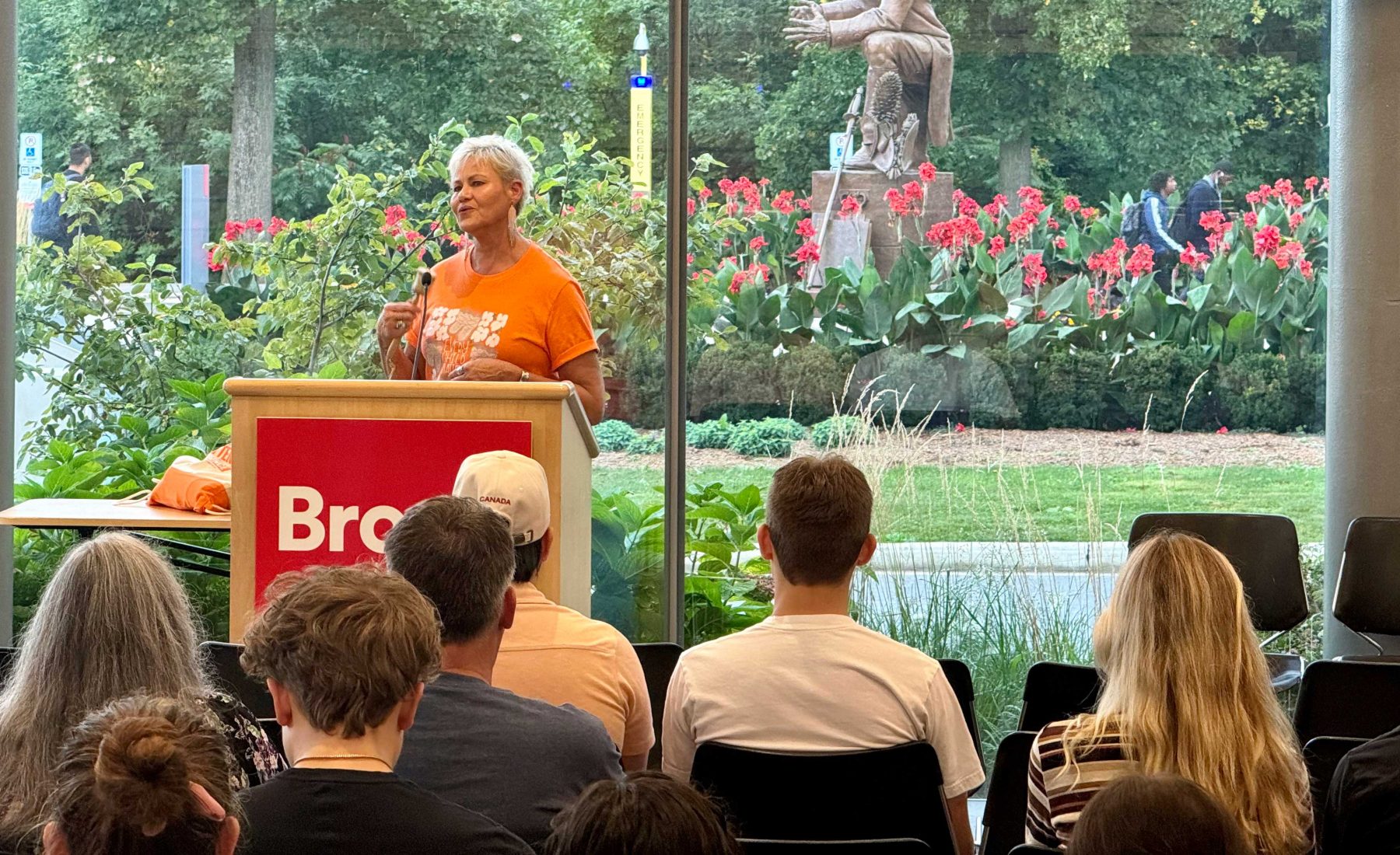 A woman wearing an orange tee shirt and standing at a Brock podium sings to an audience while shaking a traditional Indigenous instrument. 