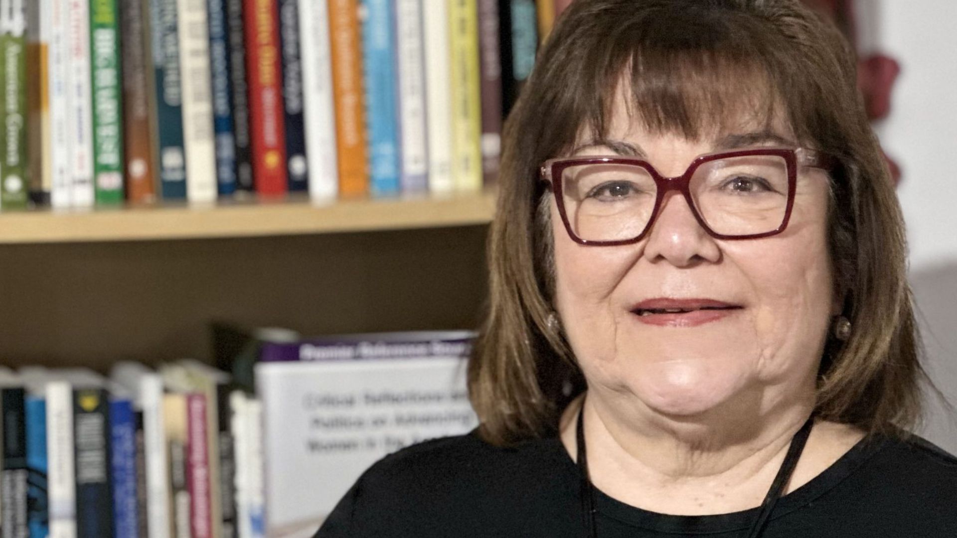 A photo of Indigenous Educational Studies prof Sheila Cote-Meek shows her sitting in front of a book shelf, smiling wearing red glasses and a black shirt. 