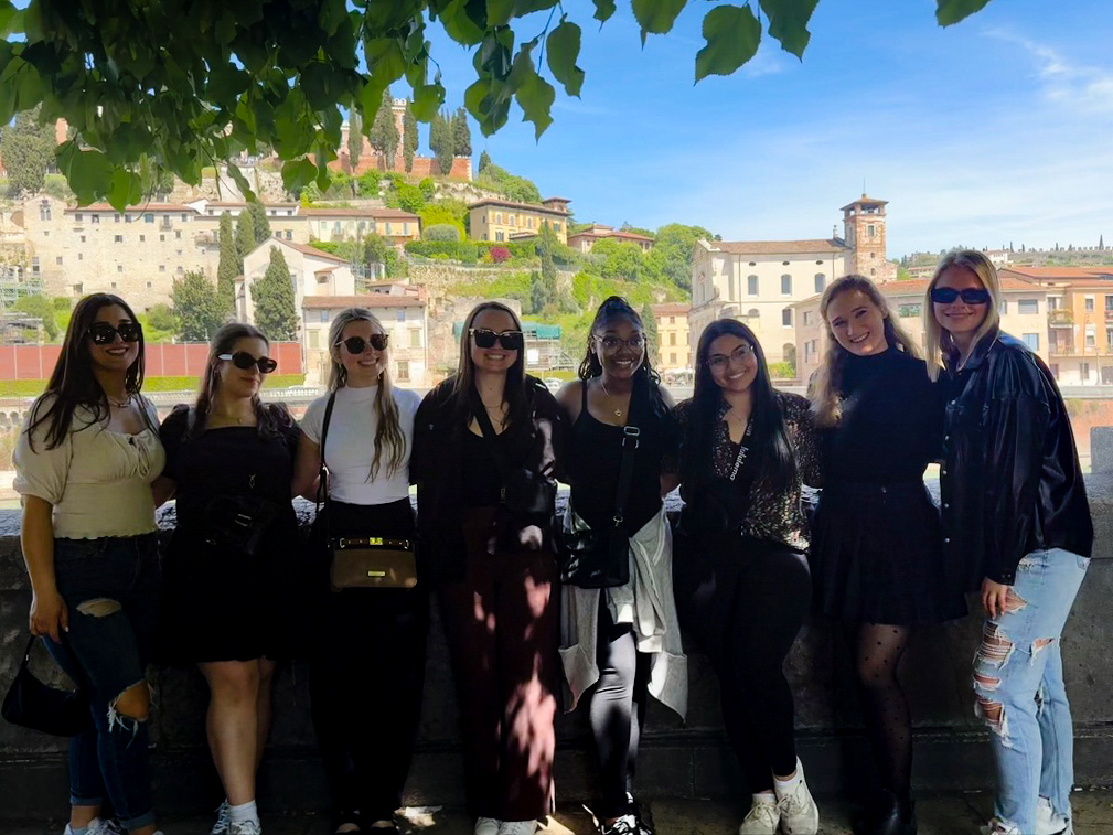 A group of women stand shoulder to shoulder in Verona with a blue sky in the background. 