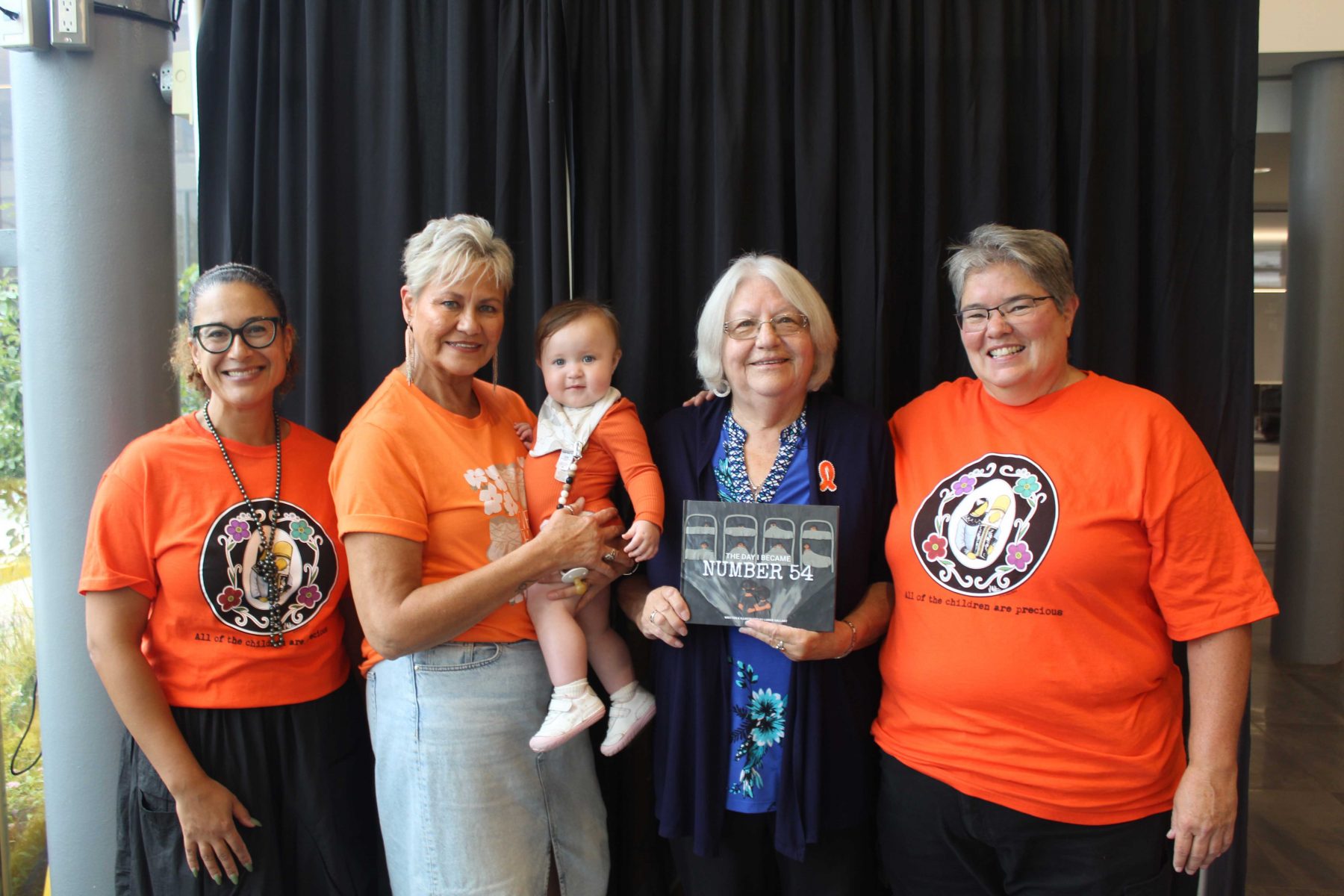 A photo shows four women standing shoulder to shoulder smiling at the camera. Three women are wearing orange shirts. One woman holds a baby in an orange onesie while Dawn Hill holds a copy of a book inspired by her life.