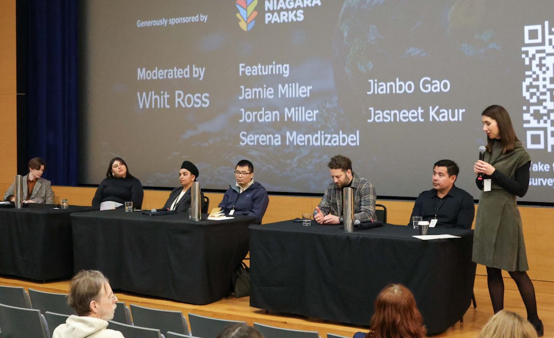 A group of people sit at a table on stage during a panel while a woman holding a microphone stands nearby. 