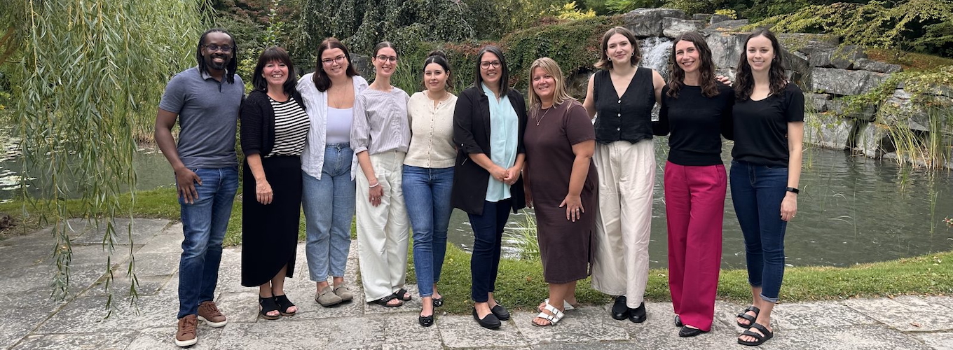 A group of people pose for a photo in front of a pond with trees and a building in the background.