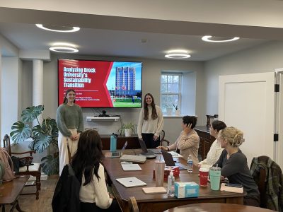 Two students delivering a presentation to a small group of people sitting around a boardroom table.