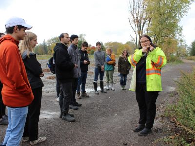 Someone in a neon yellow vest standing outside and speaking with a group of students.